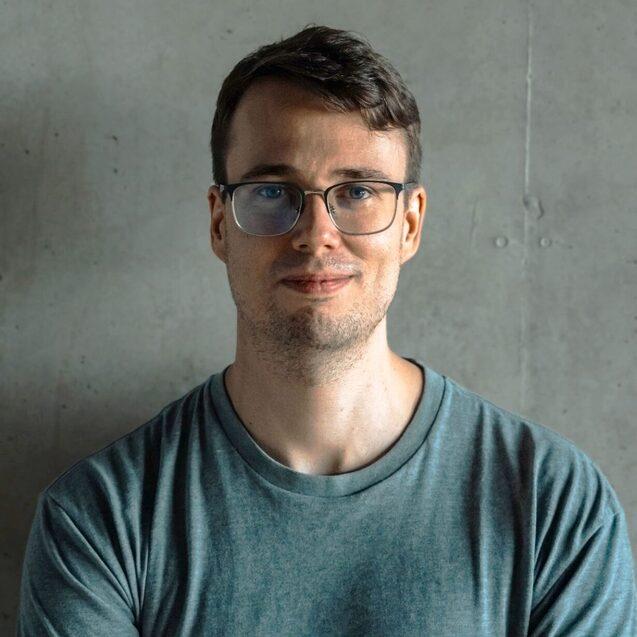 Porträt eines Mannes mit Brille, der in einem grauen T-Shirt vor einer Betonwand lächelt.