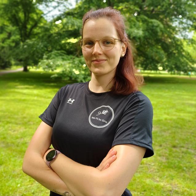 Junge Frau mit kurzen, roten Haaren und Brille, in schwarzem Sportshirt, steht in einem Park mit Grünflächen.