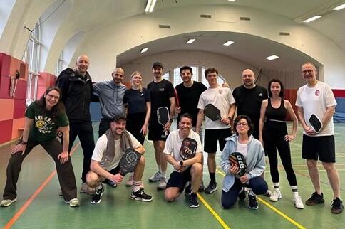 Gruppe von zwölf Personen posiert mit Paddeln in einer Sporthalle mit grüner Bodenfläche und hohen Fenstern.