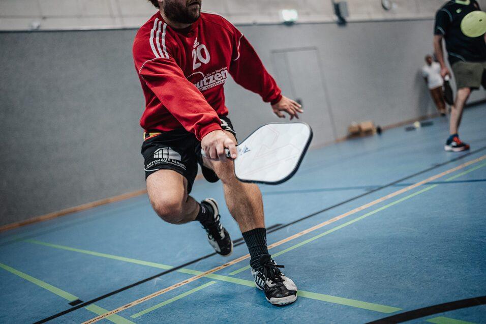 Aktiver Spieler in rotem Shirt schlägt mit einem Paddel auf einem blauen Sportboden. Hinter ihm sind weitere Personen zu sehen.