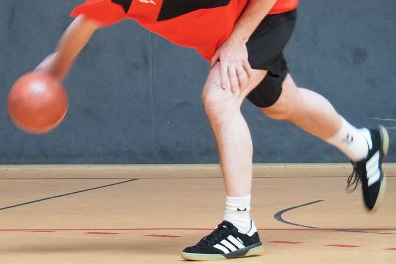 Nahaufnahme eines Spielers, der einen Basketball beim Dribbeln auf einem Holzboden bewegt. Spieler trägt sportliche Kleidung.