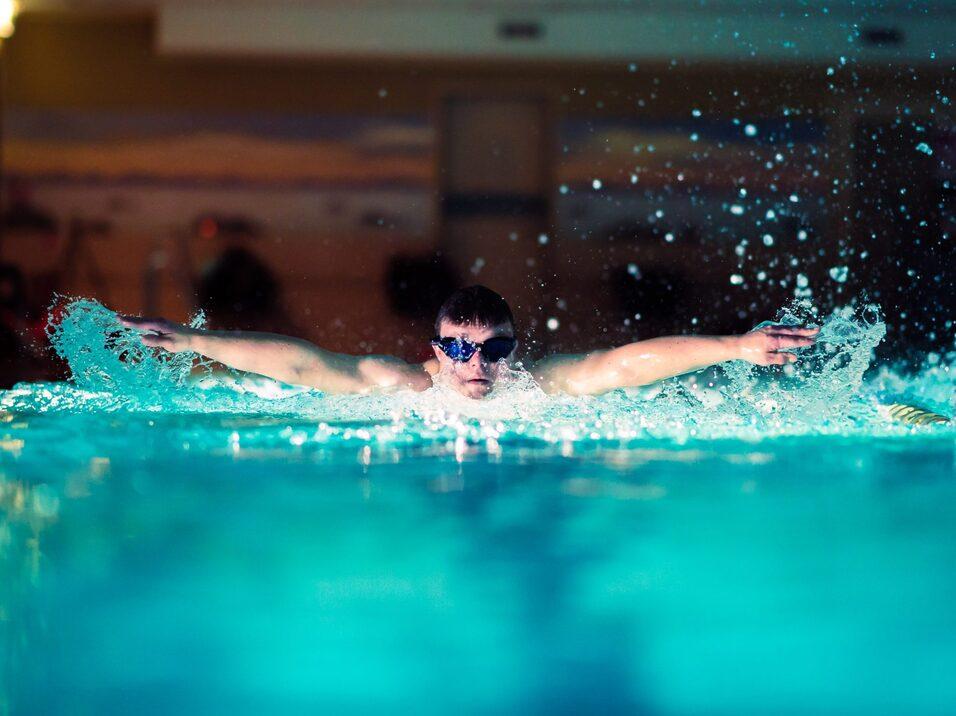 Schwimmer mit Schwimmbrille vollführt einen Kraullschlag in einem beleuchteten Pool. Spritzendes Wasser um ihn herum.