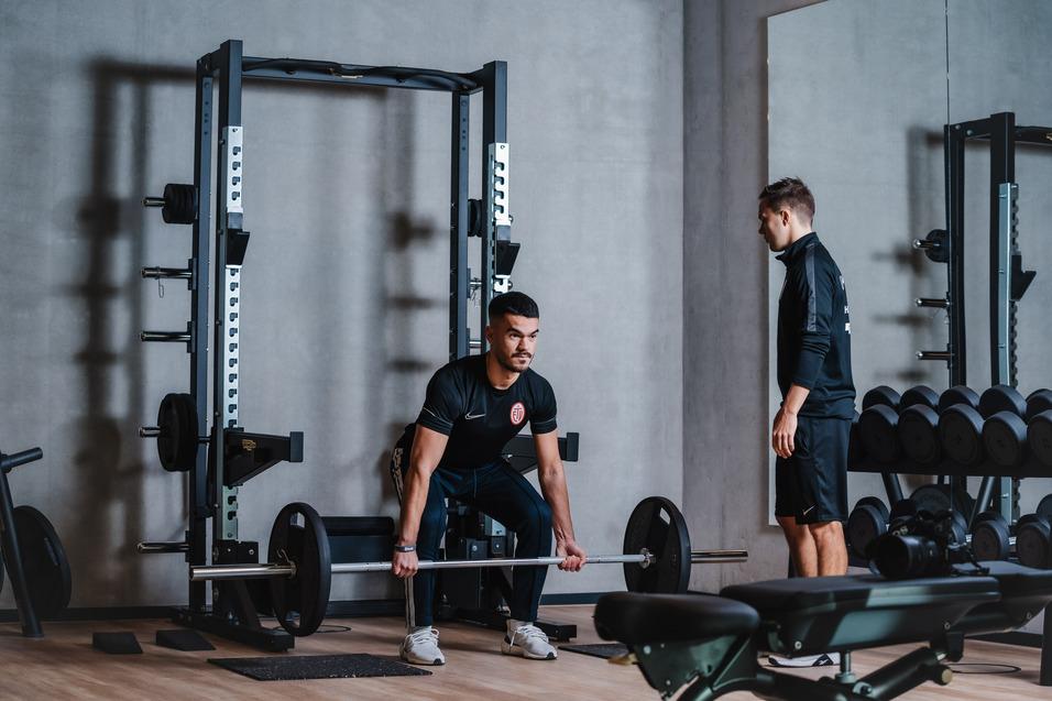 Mann hebt eine Langhantel im Fitnessstudio, während ein Trainer daneben zuschaut und Anweisungen gibt.