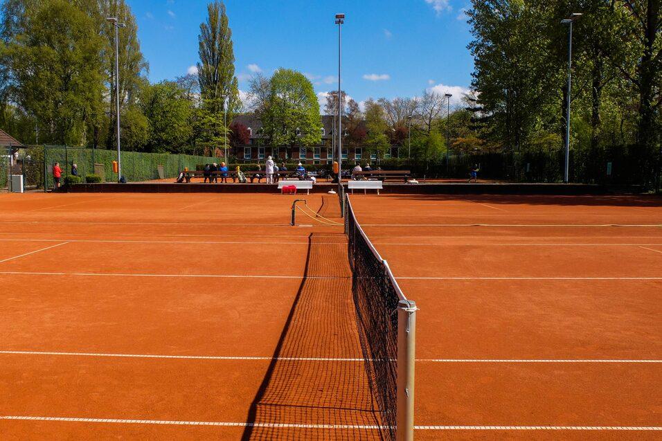 Blick auf einen Tennisplatz mit roter Erde, umgeben von Bäumen und einem offenen Bereich für Zuschauer.