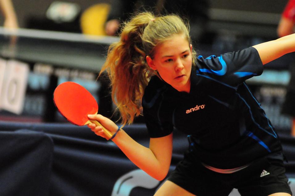 Jugendliche Spielerin schlägt mit einem roten Tischtennisschläger einen Ball während eines Wettkampfs.