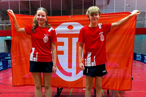 Zwei jugendliche Tischtennisspieler in roten Trikots stehen vor einer großen Flagge mit Vereinslogo.
