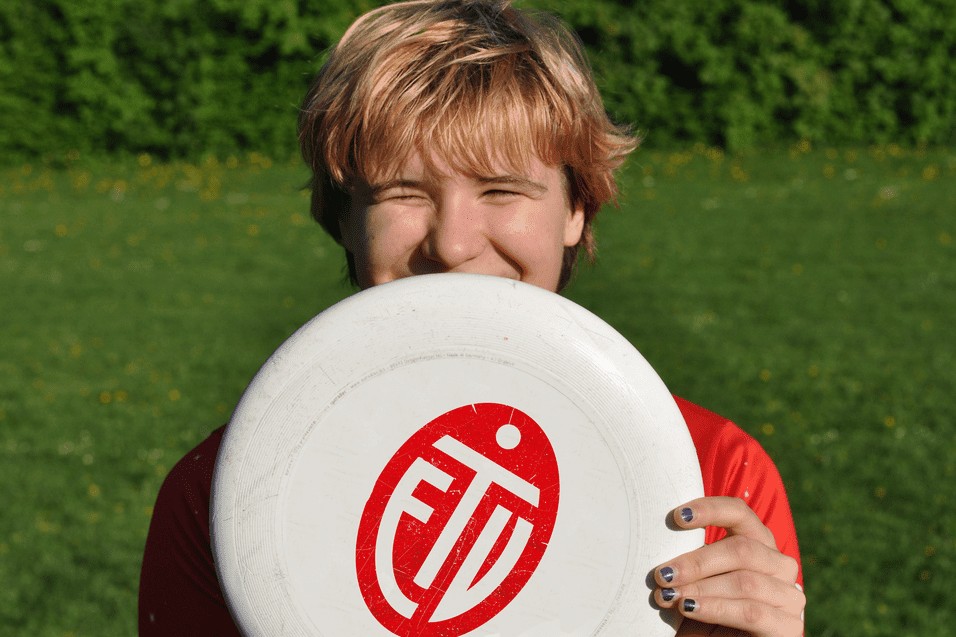 Lächelnder junger Mann hält eine Frisbee vor seinem Gesicht, auf einer grünen Wiese mit Blüten.