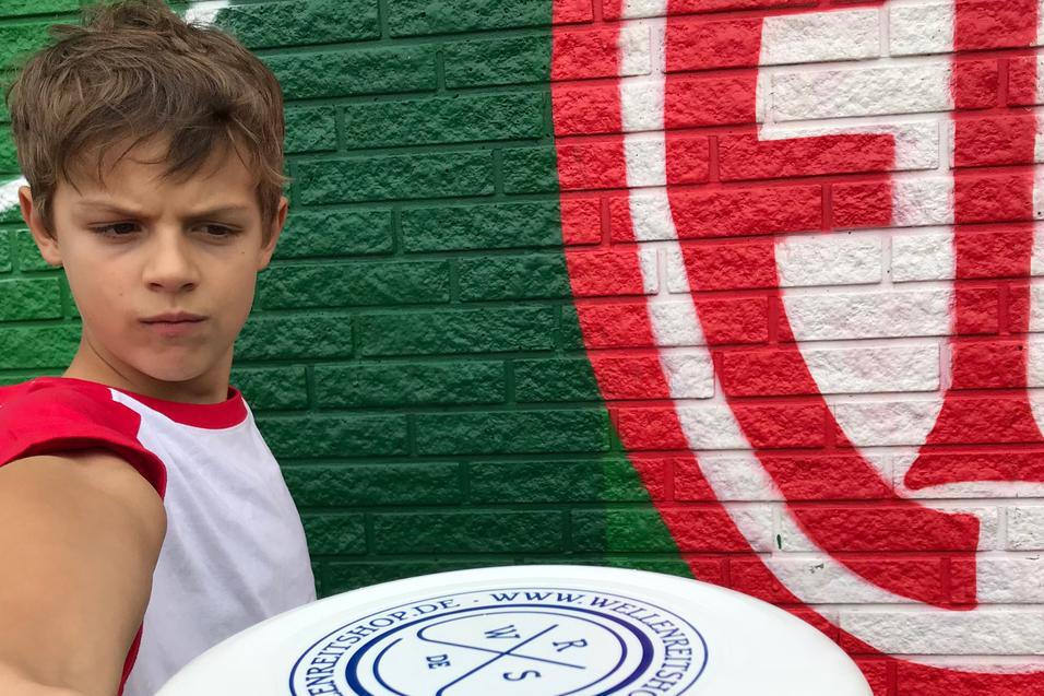 Ein Junge hält eine Frisbee in Richtung Kamera, vor einer bunten Wand mit grünen und roten Mustern.