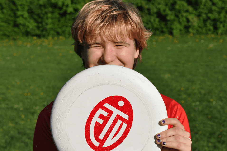 Lächelnder junger Mann hält eine Frisbee vor seinem Gesicht, auf einer grünen Wiese mit Blüten.