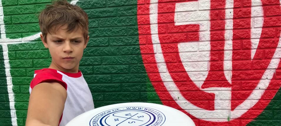 Ein Junge hält eine Frisbee in Richtung Kamera, vor einer bunten Wand mit grünen und roten Mustern.