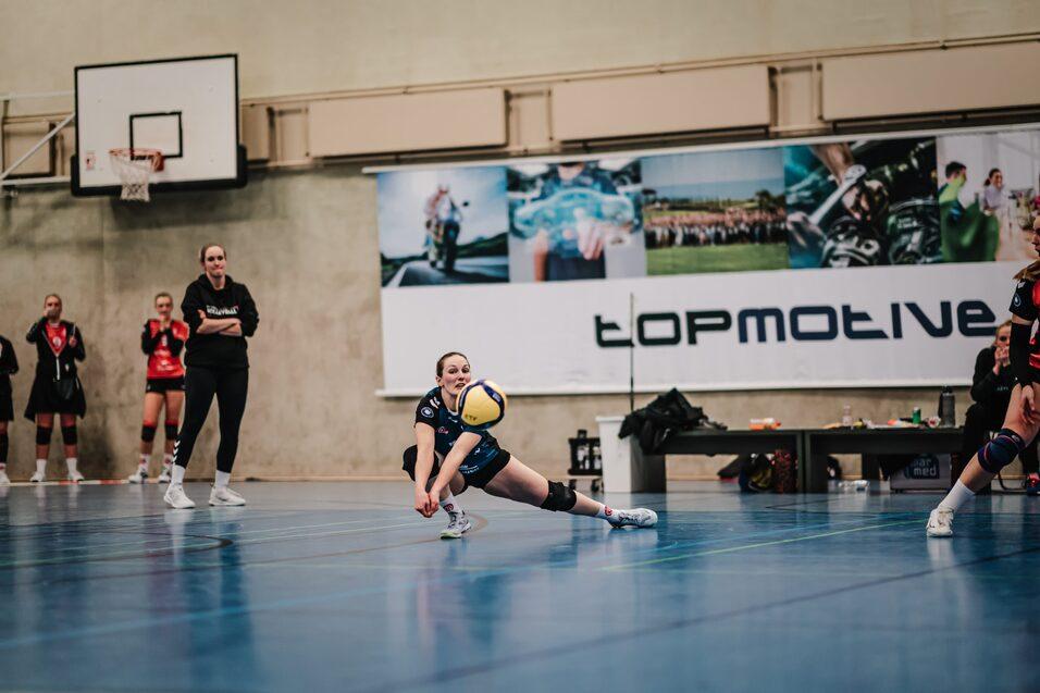 Spielerin macht einen Wurf mit einem Volleybal in einer Sporthalle, während Zuschauer im Hintergrund zuschauen.