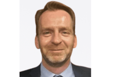 Porträt eines Mannes mit lächelndem Gesicht, tragt Anzug und Krawatte mit grauem Hintergrund.