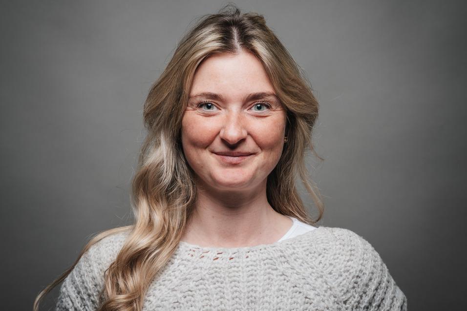 Porträt einer lächelnden Frau mit langen, welligen Haaren, die einen hellen Strickpullover trägt, vor grauem Hintergrund.