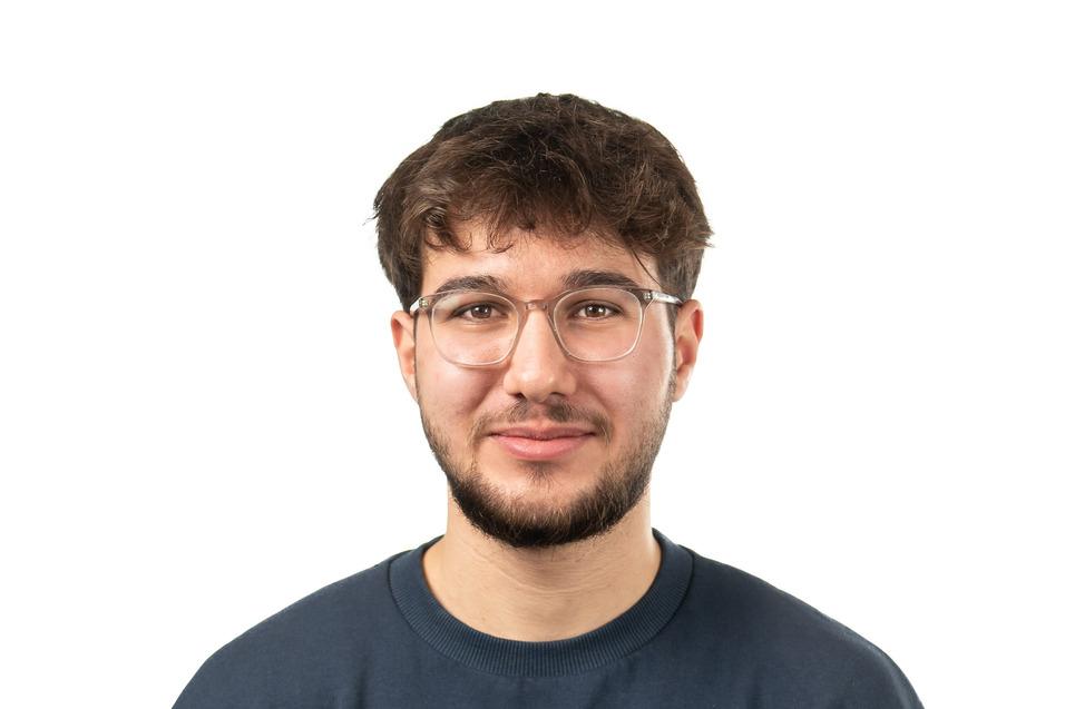 Junger Mann mit Brille und T-Shirt mit Bergmotiv steht vor neutralem Hintergrund.
