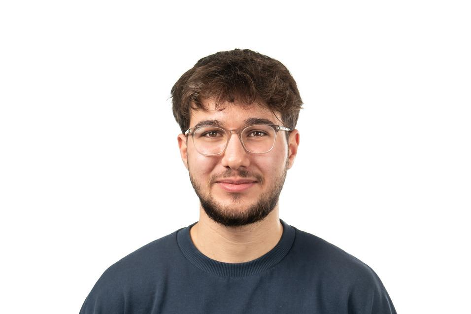 Junger Mann mit Brille und T-Shirt mit Bergmotiv steht vor neutralem Hintergrund.