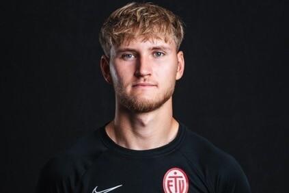 Porträt eines jungen Mannes mit blondem Haar, in einem schwarzen Sporttrikot mit Logo auf dunklem Hintergrund.