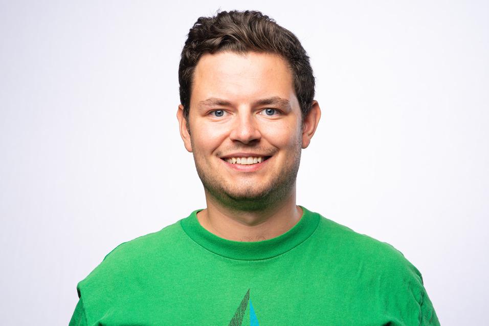 Lächelnder Mann mit kurzen, dunklen Haaren, trägt ein grünes T-Shirt mit geometrischem Muster vor hellem Hintergrund.