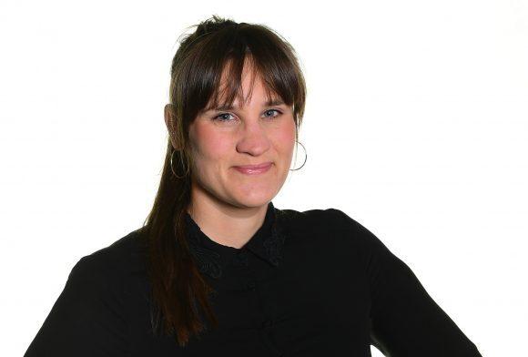 Porträt einer lächelnden Frau mit langen, braunen Haaren, die in einem schwarzen Oberteil auf einem weißen Hintergrund steht.