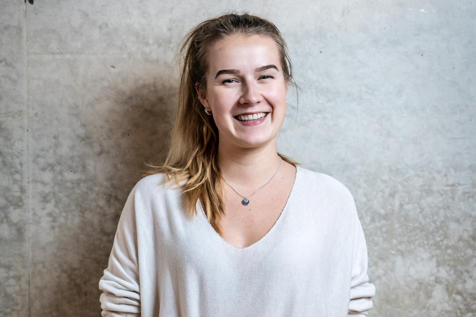 Junge Frau mit lässigem Outfit lächelt vor einer grauen Betonwand. Sie trägt einen hellen Pullover und eine dunkelblaue Hose.