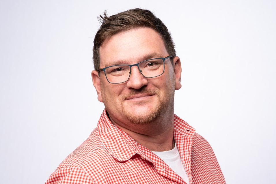 Mann mit Brille und gestreiftem Hemd lächelt, vor einem einfarbigen Hintergrund.