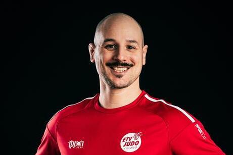 Mann mit Glatze und Schnurrbart trägt ein rotes Judoshirt und steht lächelnd mit akzentuierten Händen vor schwarzem Hintergrund.