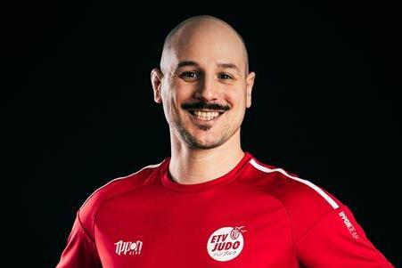 Mann mit Glatze und Schnurrbart trägt ein rotes Judoshirt und steht lächelnd mit akzentuierten Händen vor schwarzem Hintergrund.