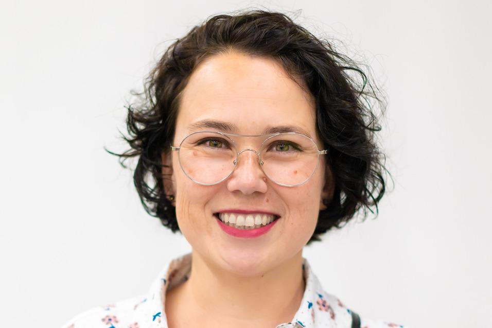 Portrait einer Frau mit lockigem Haar und Brille, die ein blumenmusterndes Hemd trägt, vor einem weißen Hintergrund.