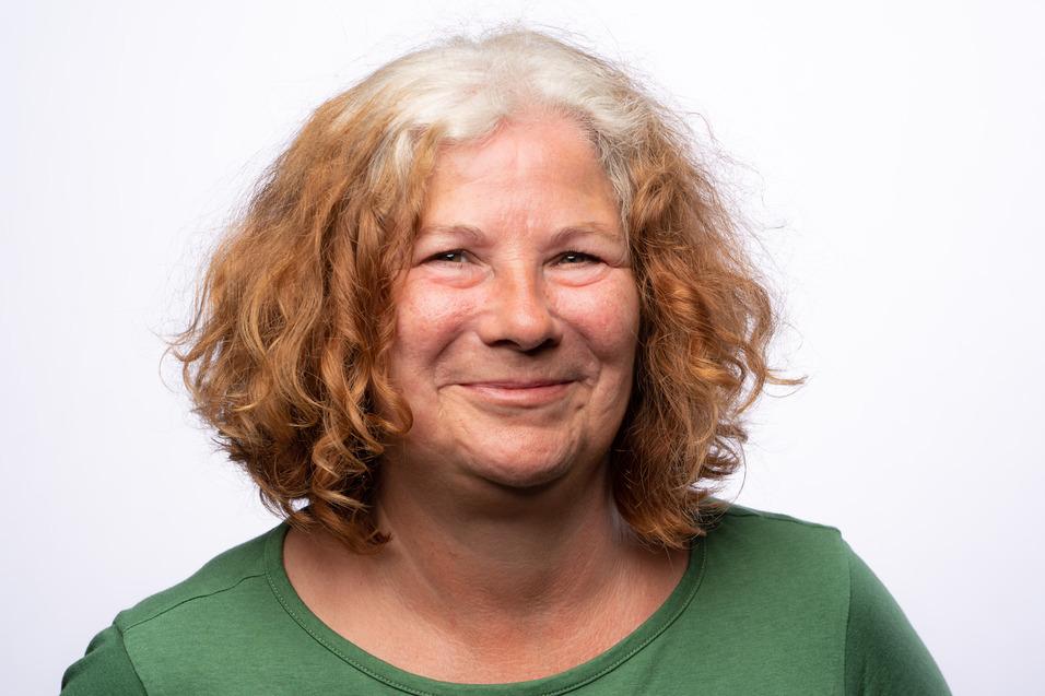 Lächelnde Frau mit lockigem, brünett-grauem Haar in einem grünen Oberteil vor hellem Hintergrund.