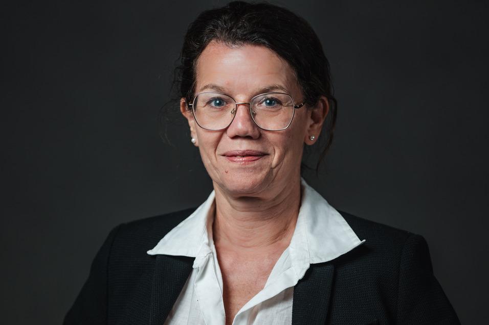 Portrait einer Frau mit Brille, in schwarzem Blazer und weißem Hemd, vor einem dunklen Hintergrund.