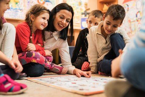 Gruppe von Kindern und einer Lehrerin sitzt im Kreis auf dem Boden und spielt ein Brettspiel in einem bunten Raum.