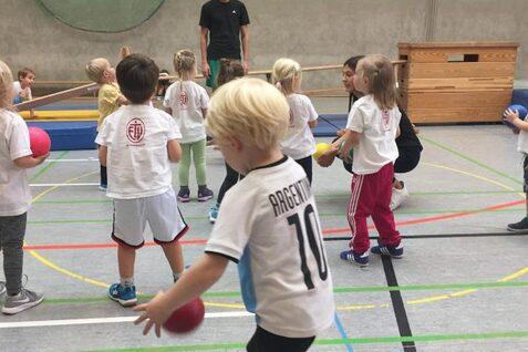 Kinder in Sportkleidung spielen mit bunten Bällen in einer Turnhalle, während sie von Erwachsenen angeleitet werden.