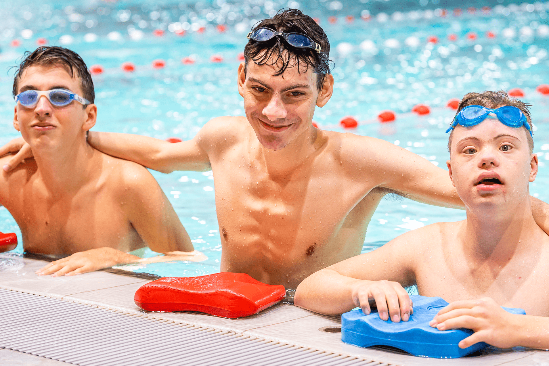 Drei Schwimmer in Badekappen und Schwimmbrillen lehnen im Wasser auf eine Kante, die Schwimmbahnen sind im Hintergrund sichtbar.