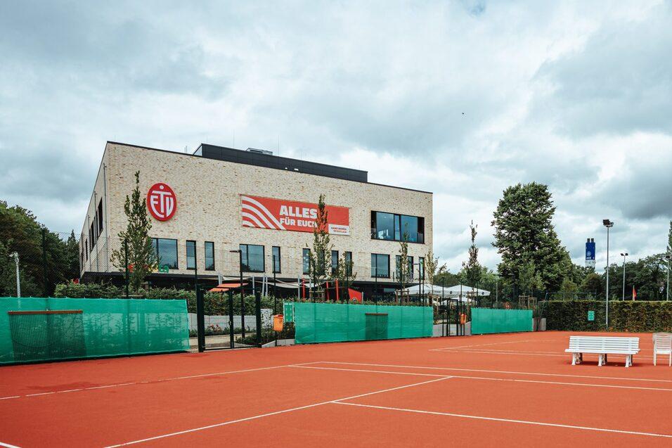 Tennisplatz mit rotem Belag und einem modernen Gebäude im Hintergrund, umgeben von grünen Netzen und Bäumen.