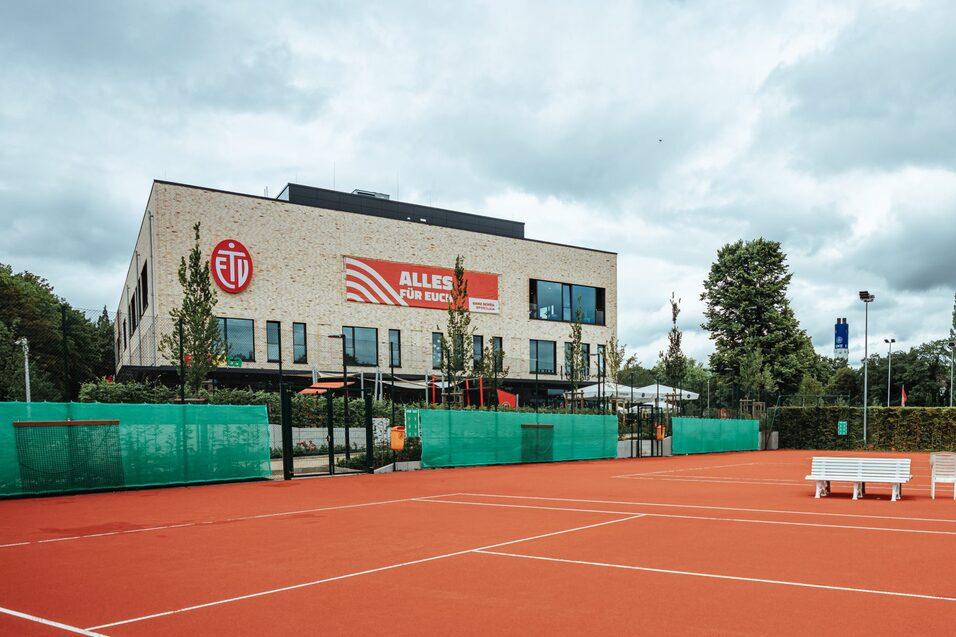 Tennisplatz mit rotem Belag und einem modernen Gebäude im Hintergrund, umgeben von grünen Netzen und Bäumen.