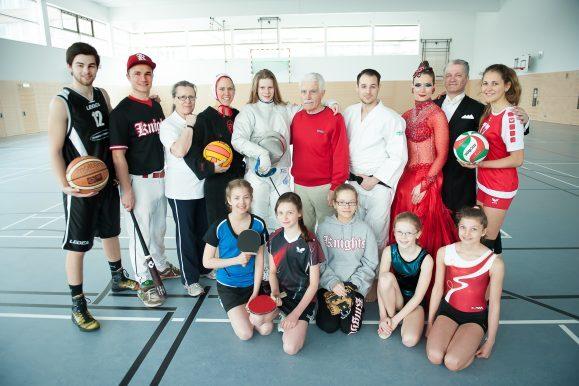 Gruppe von Sportlern in einem Sporthallen, diverse Sportgeräte wie Basketball, Tischtennisschläger und Volleyball in den Händen.