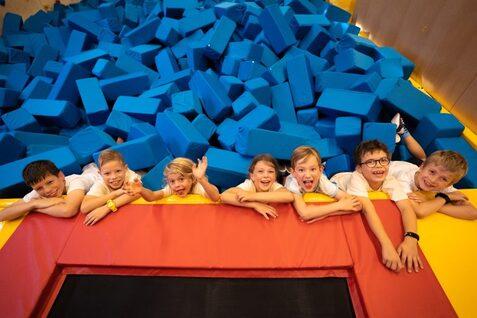 Sechs Kinder lehnen auf einer roten Trampolinrand über einem blauen Schaumstoffblock-Pool und freuen sich.