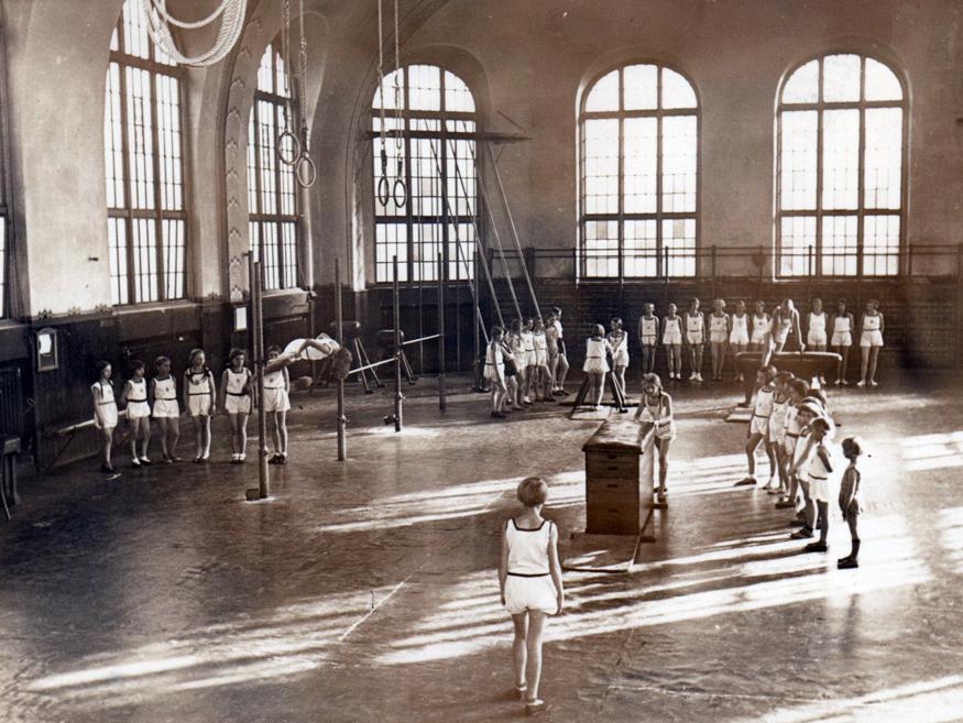 Gruppe von Turnern in einer Sporthalle, die verschiedene Übungen an Geräten ausführen, umgeben von großen Fenstern.