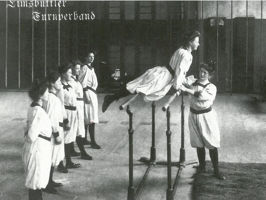 Frauen turnen an einem niedrigem Schwebebalken, beobachtet von mehreren anderen in historischen Turnanzügen.