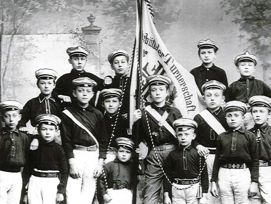 Gruppenfoto von Jungen in Uniformen mit Hüten, die eine Fahne der Turnerschaft halten, vor einer landschaftlichen Kulisse.
