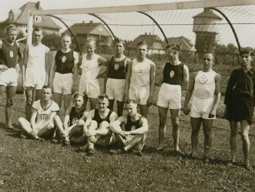 Gruppensfoto von zwölf Sportlern in Trikots auf einem Fußballfeld, umgeben von einem Tor und Gebäuden im Hintergrund.