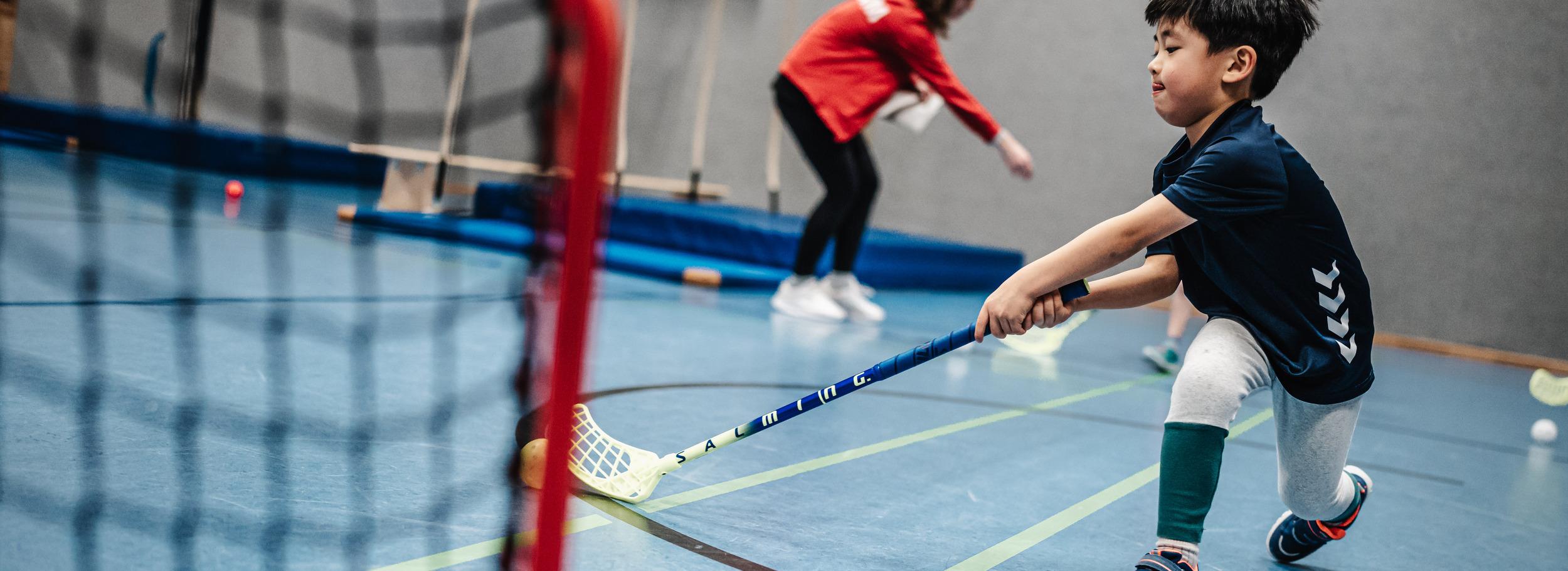 Junge mit einem Stock im Hallen-Sportraum, während er auf ein Tor zielt. Im Hintergrund eine Trainerin.