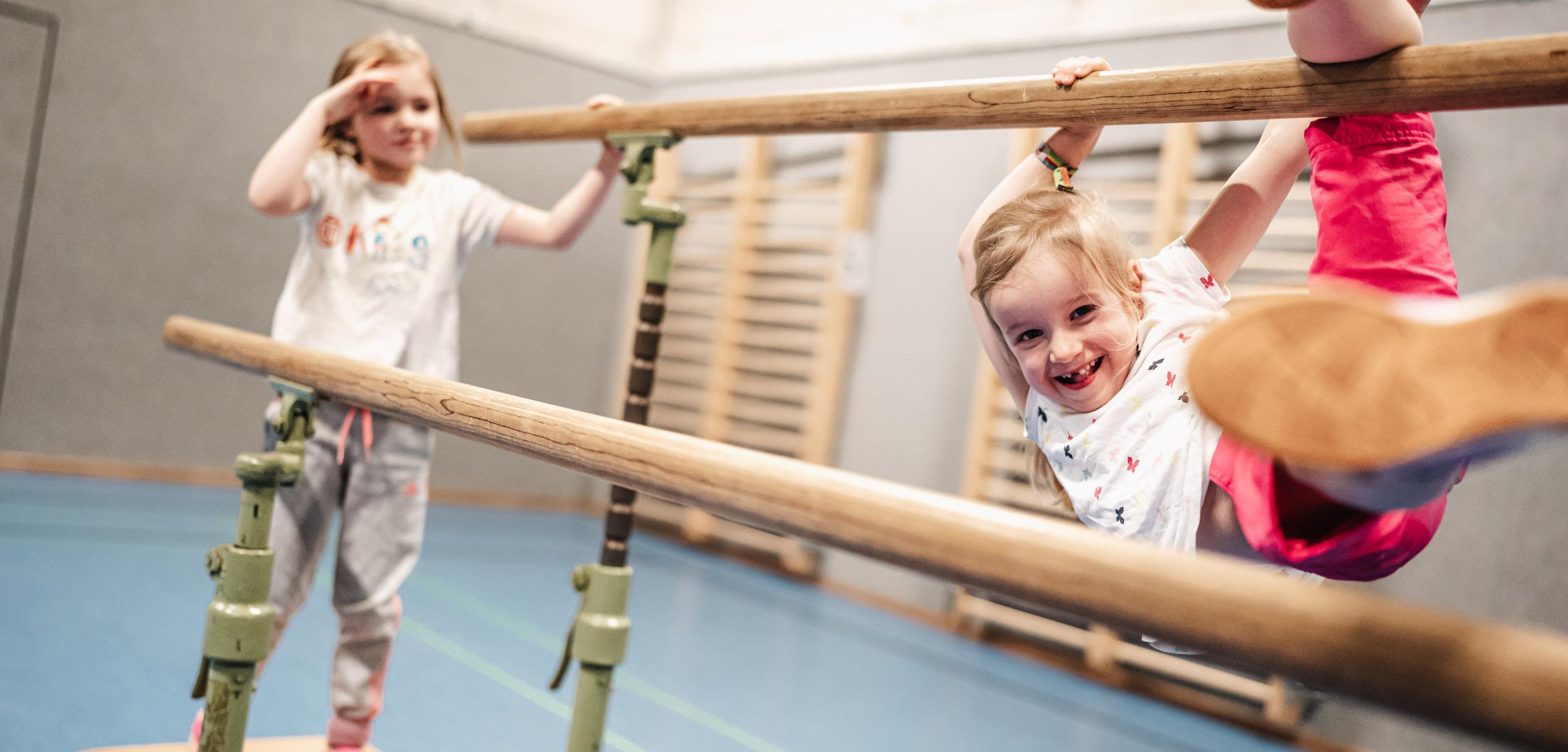 Zwei Kinder spielen in einer Sporthalle an einem Barren, eines hängt lächelnd, das andere steht daneben.