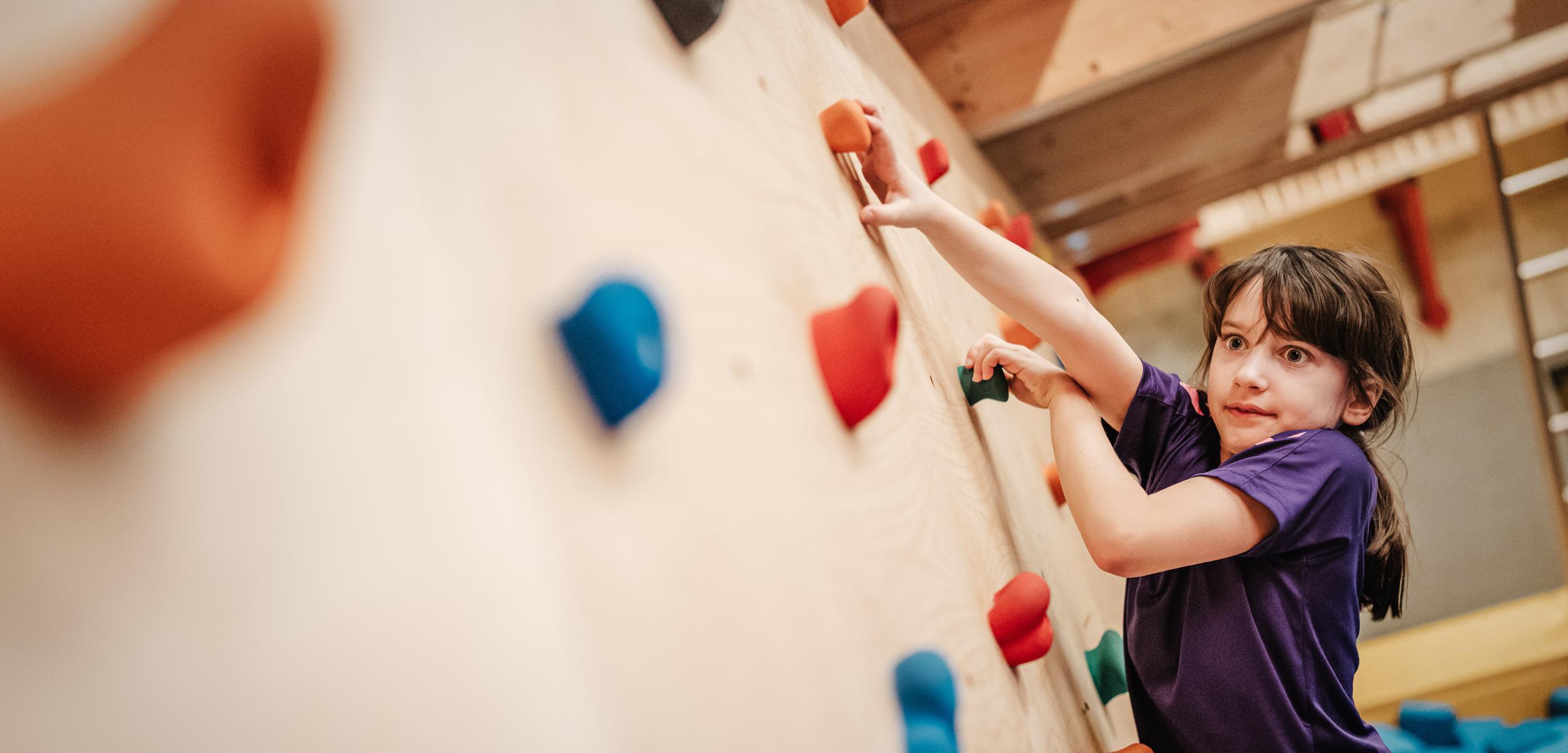 Mädchen klettert an einer bunten Kletterwand mit verschiedenen Griffen in einer Turnhalle.