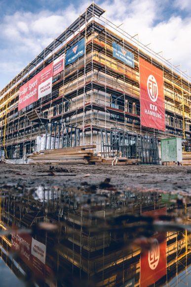 Baustelle eines mehrstöckigen Gebäudes mit einem Gerüst und Werbebannern, reflektiert in einer Pfütze im Vordergrund.