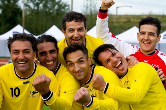 Sechs Fußballspieler in gelben Trikots lächeln und posieren fröhlich miteinander auf einem Sportplatz.