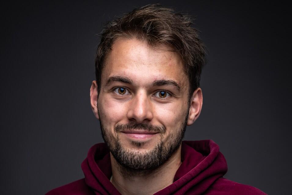 Porträt eines lächelnden Mannes mit kurzen braunen Haaren, der einen roten Hoodie trägt, vor dunklem Hintergrund.