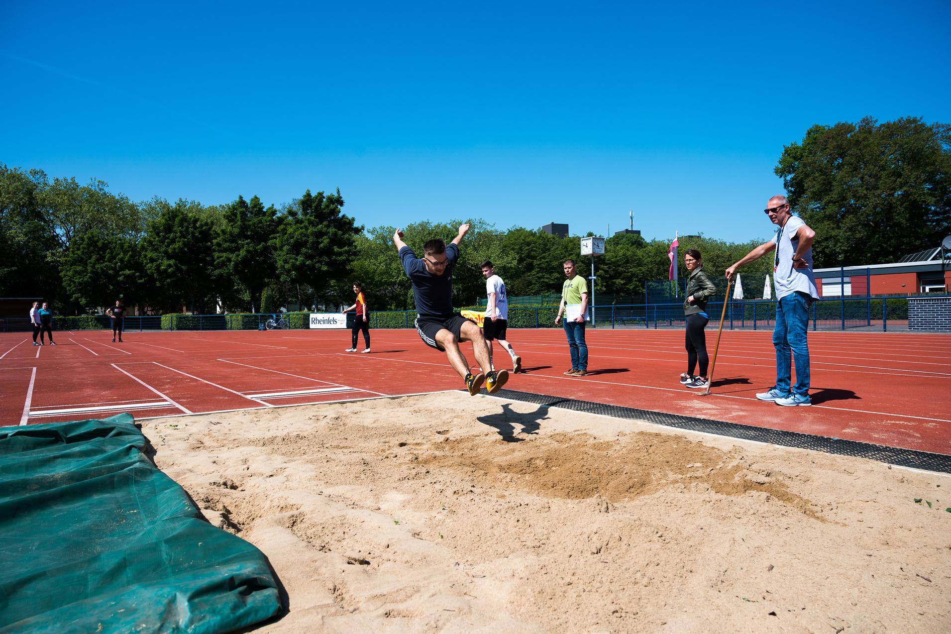 Sprinter springt vom Absprungbrett in die Sandschicht einer Weitsprunganlage, während Zuschauer am Rand stehen.