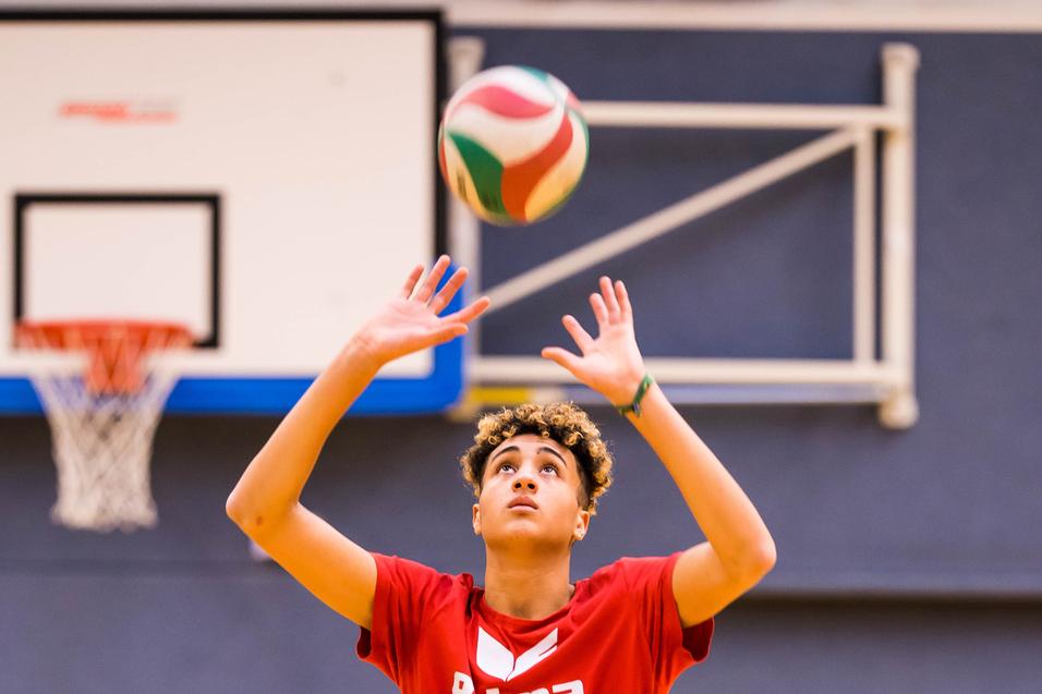 Junger Volleyballspieler hebt den Ball zum Spielen über den Kopf, in einer Sporthalle mit Basketballkorb im Hintergrund.