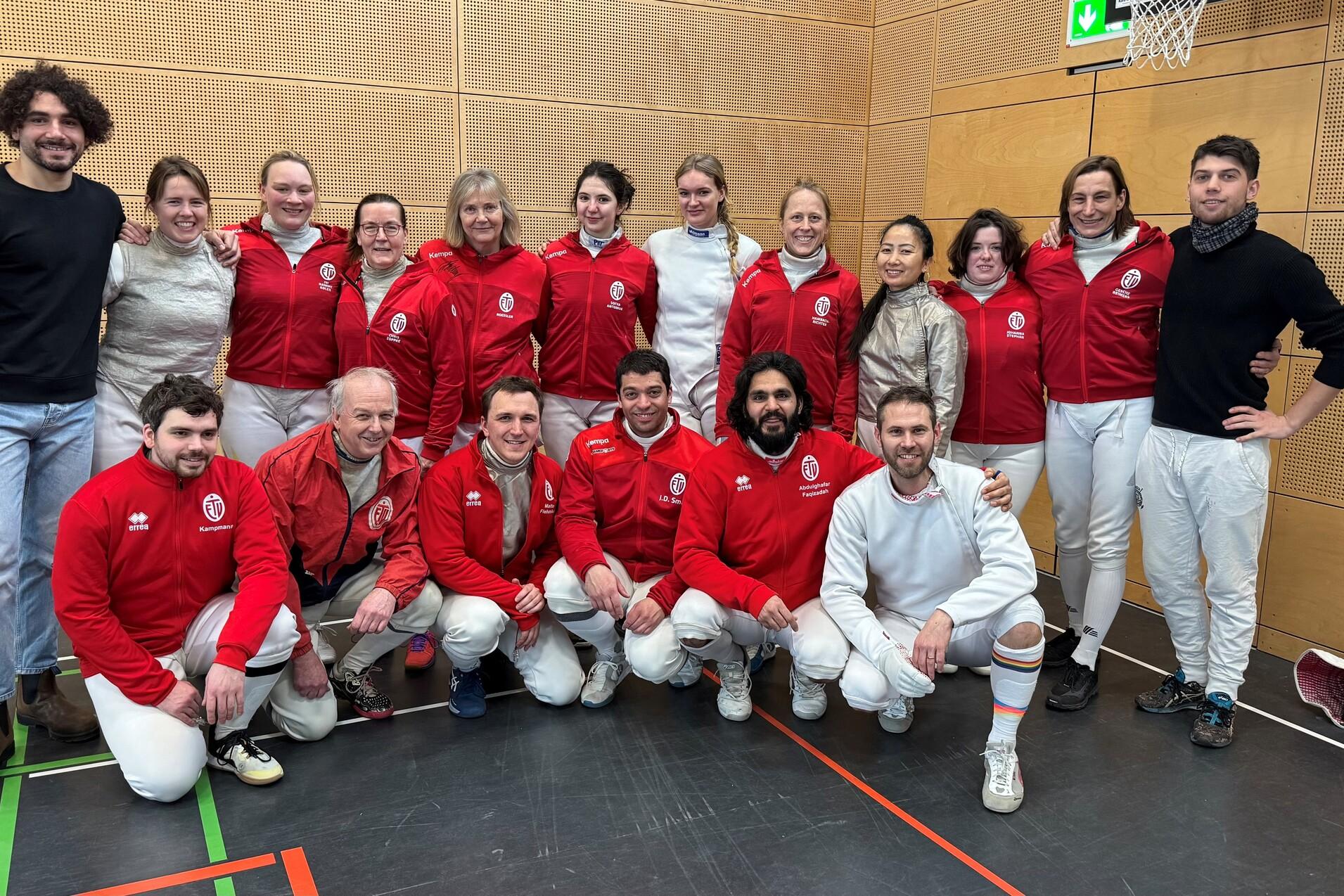 Gruppenfoto von 15 Personen in roten Trainingsjacken und weißen Sportsachen in einer Turnhalle.