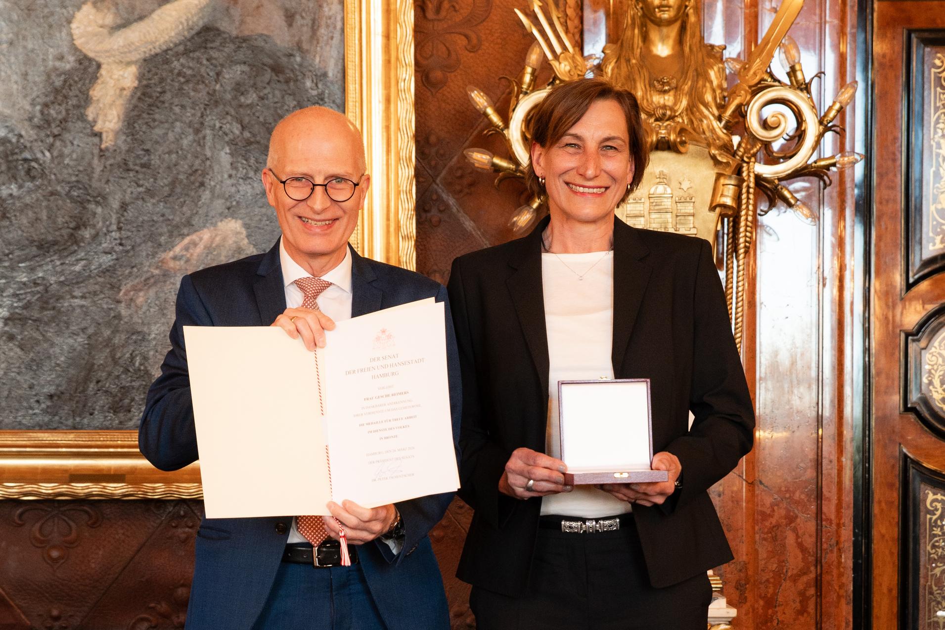 Zwei Personen in formeller Kleidung halten eine Urkunde und eine Auszeichnung in einem prunkvollen Raum.